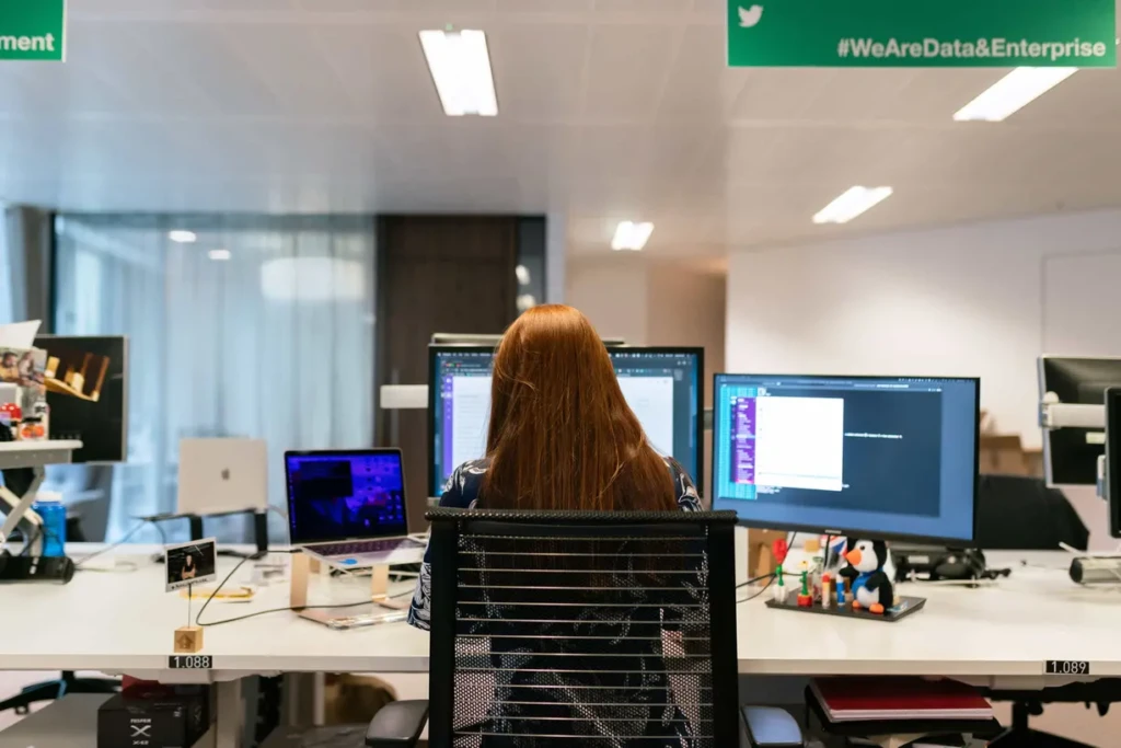 A woman sitting at a desk, focused on two computer screens, working diligently.